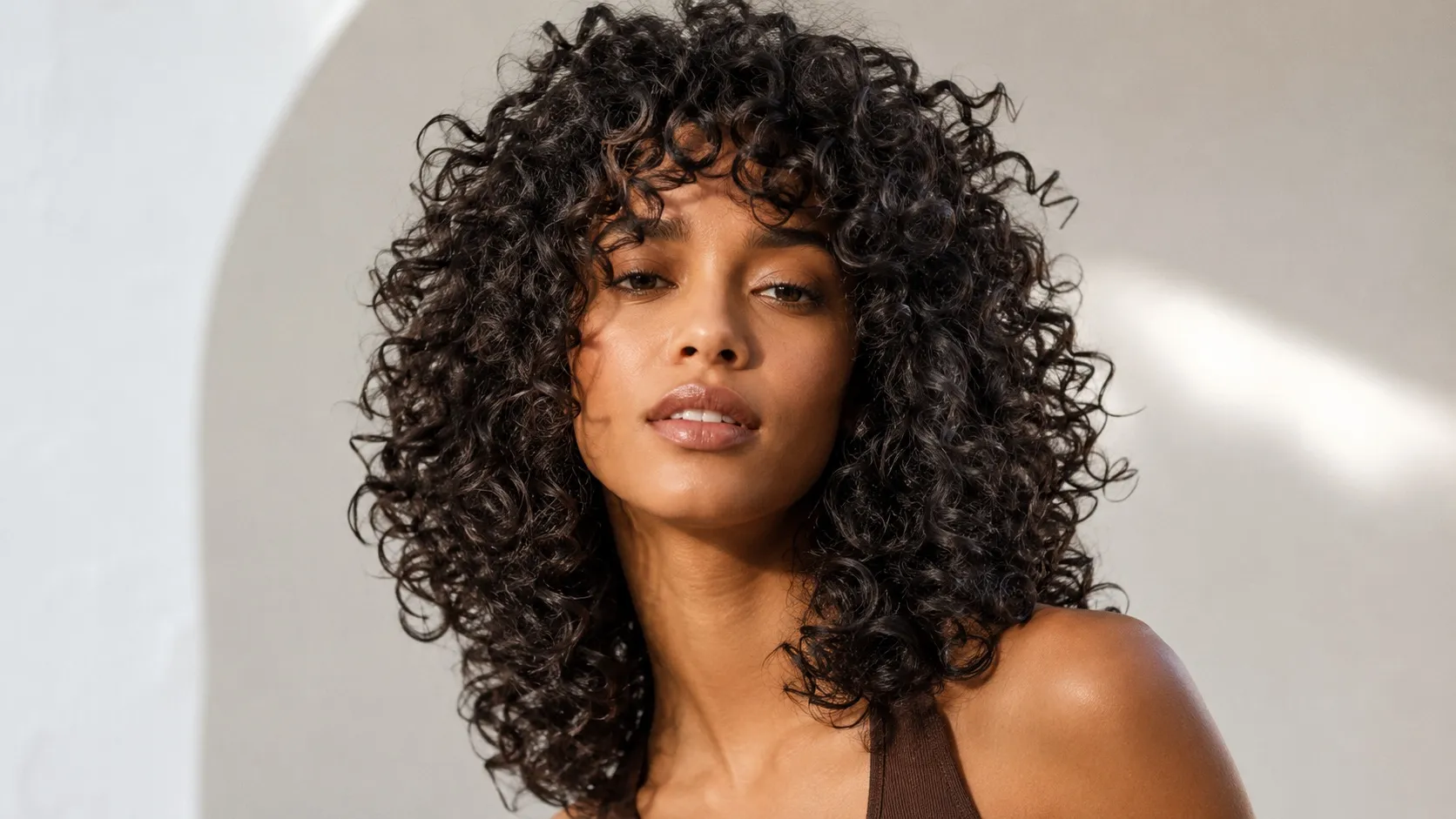 A woman with a voluminous curly shag haircut featuring defined dark curls, layered texture, and natural movement. The hairstyle has soft, bouncy curls with face-framing layers and a modern editorial look.
