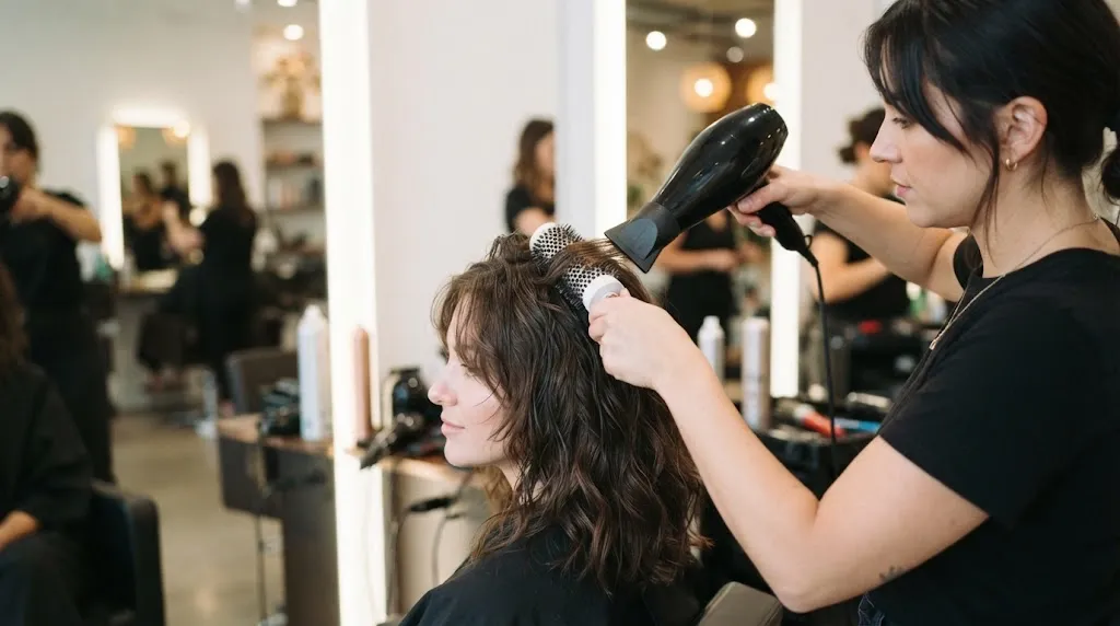 A professional stylist uses a round brush and blow-dryer to smooth a stubborn cowlick on a client's hair in a modern salon.